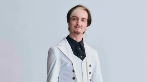 Bafta/Hollie Fernando Stanley Baxton wears a white suit jacket over a black shirt. He has dark hair with a blonde fringe. 