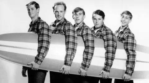 Getty Images The Beach Boys pose for a portrait with a surfboard in August 1962 in Los Angeles, California. (L-R) Brian Wilson, Mike Love, Dennis Wilson, Carl Wilson, David Marks. They are wearing matching checked shirts buttoned up over white t-shirts, and jeans.