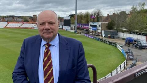 Gary Hoffman in a jacket and tie at a cricket groundnds in a cricket ground