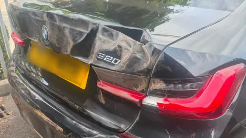 Charlotte Baker The back of a black BMW which has been heavily damaged. The red rear lights are smashed and the body has been deformed. 