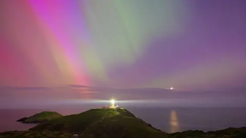 Getty Images A lighthouse's light flashes towards the camera, and is surrounded by a purple horizon of sea and sky. Downward streaks of green are among the purple sky