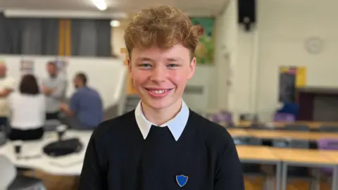 Smiling Aleksander has short, wavy hair and is wearing a school uniform. He is in a classroom