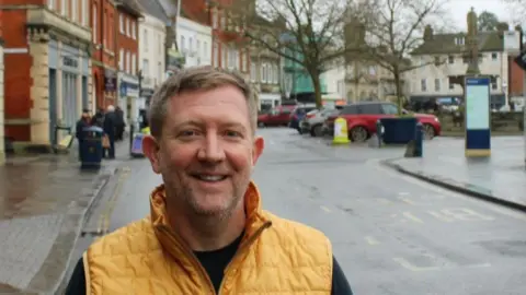 Ben Reed A man in a yellow gilet stood in a town centre with parked cars and buildings visible behind him