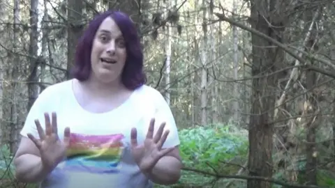 YouTube Zoe Watts stands in a forest surrounded by trees. She is wearing a white t-shirt with a rainbow flag on it.