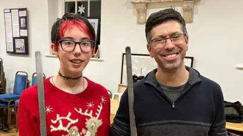Two people standing inside a hall holding round bladed swords. They are both smiling.