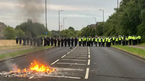Police line in front of protesters with a small fire burning in the foreground on the road at Manvers Way, Rotherham.