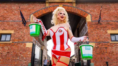 View of a drag queen wearing a full blonde wig and dressed in a red and white striped corset with a logo of Sunderland FC on the left breast, white fingerless gloves and red fishnet stockings. She is holding two fundraising buckets and standing in front of the open arch of a high brick building.