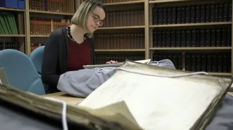 Alexandra Medcalf The image shows a woman with medium length blonde hair and spectacles sitting at a desk in an archive room, surrounded by shelves filled with large, old books. She is examining historical documents laid out on the table, some of which are propped on padded supports to protect the pages. 