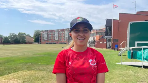 Tom Williams/BBC Firooza Amiri smiles at the camera while standing next to the cricket pitch. She wears the red T-Shirt of her kit, and a black baseball cap. Her fair hair is tied back. 