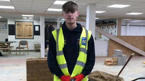 Heath is standing in a white workshop with bricks and a table and chair in the background. He has short brown hair and is looking into the camera, standing with his hands folded in front of him. He is wearing a black long-sleeved top with a high-vis sleeveless jacket over the top and red gloves