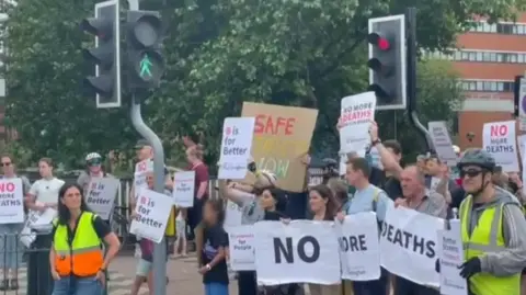 Campaigners are holding placards that call for "no more deaths" and calls for road safety improvements. They are stood by some traffic lights. A 'green man' light is showing on the lights for pedestrians to cross. 