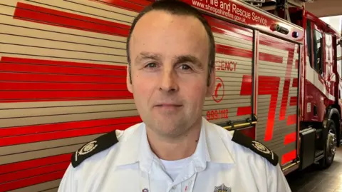 A close up of the face of a man with brown hair, in a white shirt with black epaulettes.