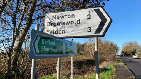 Alex Pope/BBC Two road signs, by a busy road. One is white with black writing on, the other is green. There are trees, a field to the left and cars on the road to the right. 