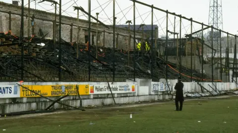 Charred remains of a football stand, strewn with tape. A single police officer looks on.
