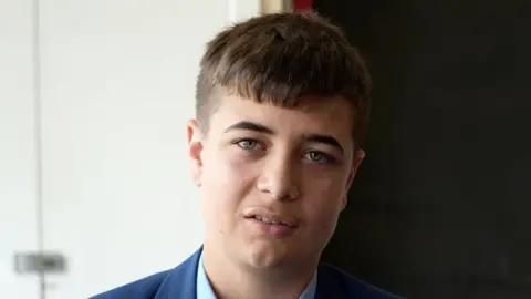 Shaun Whitmore/BBC A head and shoulders image of a young man. He is wearing a navy blazer over a smart shirt. He is looking into the camera.