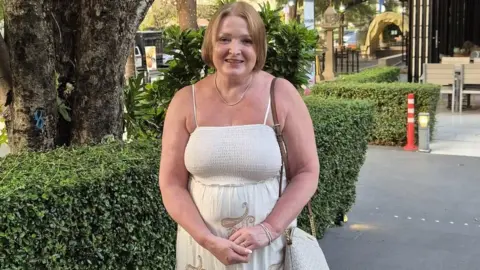 Julie Osgood, pictured in her late 50s, wearing a sleeveless white dress and with light blonde hair cut into a bob, stands in front of a green hedge and smiles at the camera.