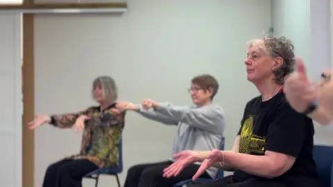 Three women sitting on chairs taking part in a movement class. The two in the background of the photo have their arms extended in front of them performing wrist circles. The woman in the foreground has her arms bent and hands open with fingers pointing down to the floor. The women at the front of the photo has short grey hair and is wearing a black t-shirt with a yellow print.