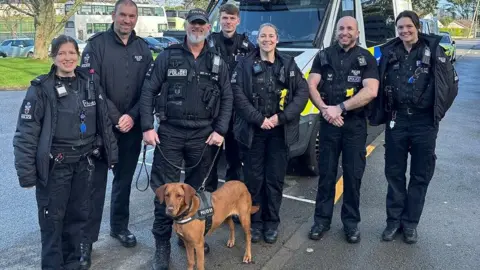Devon and Cornwall Police Seven police officers in uniform stand behind drugs dog Skye, a brown labrador type dog. They are all smiling. Behind them is a police van.
