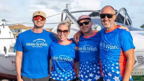 Nicky Allen Four people in light blue t-shirts with orange trim around the collars stand in front of a white boat smiling at the camera. All are wearing sunglasses. 