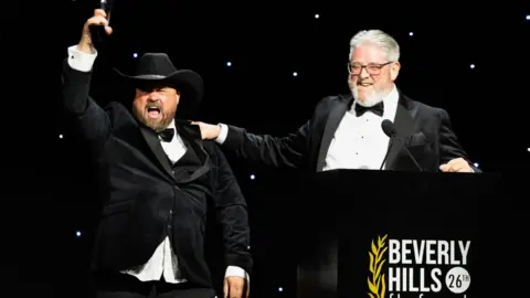 Tim Edwards Comedian John May, wearing a black tuxedo and cowboy hat, hold up an award on stage while Tim Edwards, also wearing a black tuxedo, places his hand on May's shoulder.