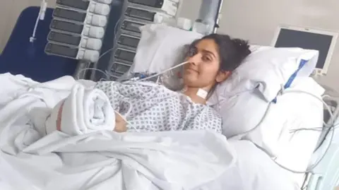 Family handout/NHS Blood and Transplant A young woman lies in a hospital bed. She has dark hair and is wearing a hospital gown. She has a tube coming out of her nose. She is looking at the camera. 