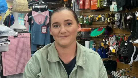 A woman stands in a sports shop. She is wearing a green jacket and a black shirt. Behind her is a collection of different sports apparel.