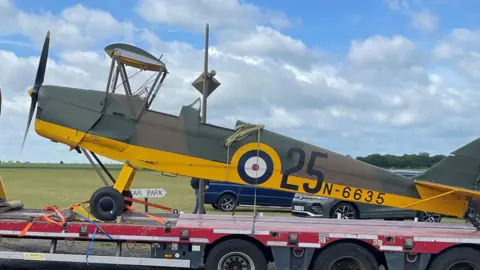 Tiger Moth N6635 on a lorry trailer. t has a grey body with a bright yellow underbelly but its two wings are missing. It has 25 and N-6635 written on its side. Behind it can be seen a field and sky.