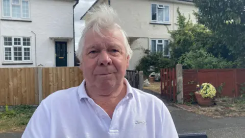 Zac Sherratt/BBC Man with white hair wearing white polo shirt looking into the camera. He is stood in front of terraced houses on a pavement