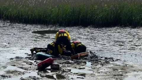 Mud with puddles on the surface with three RNLI crew members crouched together on the mud balanced on a stretcher to the right and a paddleboard to the left.