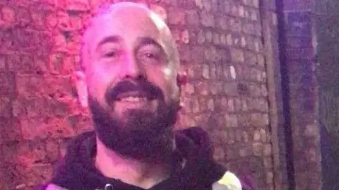 Donovan Travis Dineen-Whitham smiles at the camera. He is bald with a brown beard. He is standing in front of a wall.