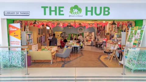 Patrick Hoelscher A wide shot of The Hub community storefront in a shopping centre, featuring the Library of Things and Greener and Cleaner logos. The open-plan interior features communal tables, a book corner, and people engaging in community activities.