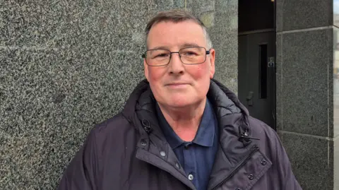 A man wearing a dark jacket, with glasses and short, dark and greying hair. He is standing outside. 