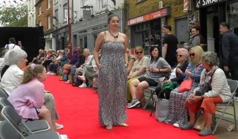 Discover Daventry Woman in blue and white strapless dress, with gold sandals and handbag, standing on a red carpet. She has dark hair, tied in a bun. A black stage is visible behind her. There are spectators in chairs on both sides of the carpet, with shops behind.