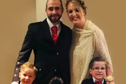 Shelley Martin Barry Shelley, and their two children at a wedding - Barry is dressed in a suit with kilt, Shelley is in a white dress and both boys are dressed formally, with black jackets, white shirts and red ties.