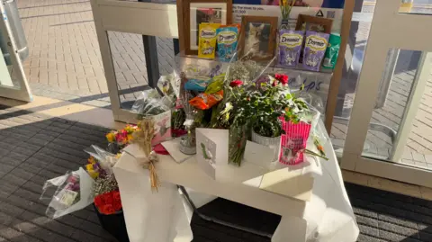 George King/BBC A shrine to Oscar the cat. Flowers, cat treats and cards can be seen placed on top of and next to a white table in the entrance of Tesco