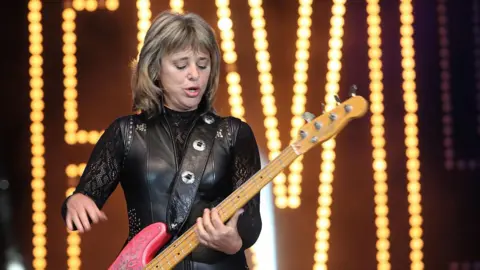 PA Media Suzi Quatro wearing a black leather outfit while playing a pink guitar on stage. She is looking down at her guitar. 