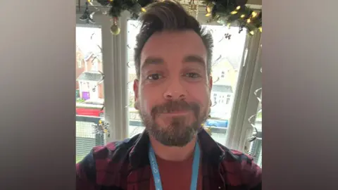 Supplied John has dark hair in a quiff and a dark beard. He is wearing a red and black checked shirt and a light blue lanyard with Claire House written on it in white writing.