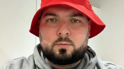 Football fan Will Grashoff has short dark hair and beard and is wearing a red hat and grey top