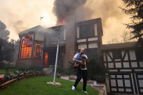 David Swanson/Reuters Seseorang yang membawa bendera AS setelah melepasnya dari tiang bendera di taman mereka berjalan menjauh dari rumah yang terbakar di Eaton Fire di Altadena pada hari Rabu.