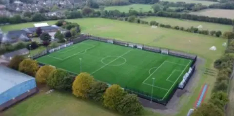 A birds-eye view of a 3G pitch nestled among green fields and trees. 