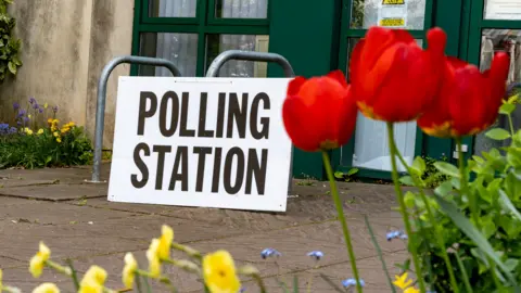 A large sign saying "POLLING STATION" in black capital letters on a white background leans against bike railings outside a building with glass doors and green door frames. In the foreground and to the left we see a selection of spring flowers including yellow daffodils, blue forget-me-nots, bluebells and red tulips. 