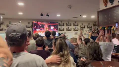 Oxford Downs Cricket Club A view of supporters cheering the choir's winning moment at the Oxford Downs Cricket Club. They are clapping and recording the TV's broadcast.