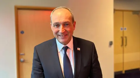 A head and shoulder shot of a man who's looking into the camera and smiling. He's inside and there are two doors behind him to his left and right. He's wearing a dark blue blazer with a white shirt underneath and a blue tie. He has a small white and red badge of the Jersey flag on his left lapel.