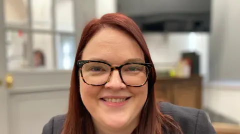 Woman wearing black jacket and glasses. She has shoulder-length brown hair.