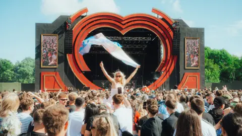 A crowd is gathered in front of a stage on a field. The stage is shaped like a red heart. A woman is on someone's shoulder facing the camera and waving a flag.