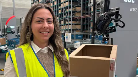 Diana Kovacs looking down the camera, standing inside a warehouse. She is wearing a yellow, high visibility gilet, over a while shirt and light brown knitted tank top. She is smiling, showing her teeth. He long, wavy brown hair falls over one of her shoulders on the right of the image. She is holding a pair of trainers, wrapped in a cellophane bag. Next to her is a cardboard box, at about chest height. The box is sitting on top of a small robot, with another robot queuing up behind, also carrying a box. Further in the background are other workstations and shelving holding other boxes.
