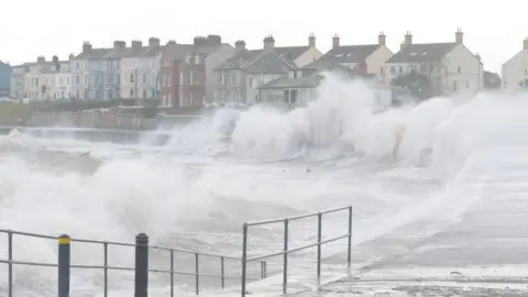 Large waves battering against the coast with multi-coloured buildings in the distance.