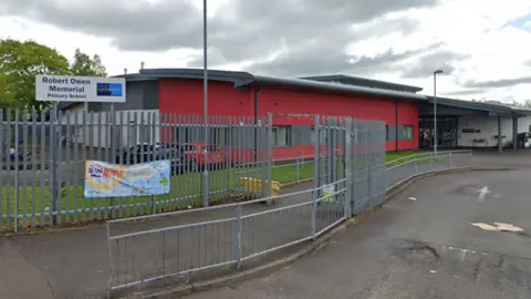 A school, with buildings coloured red and white. Gates lead into both the school and a car park nearby.
