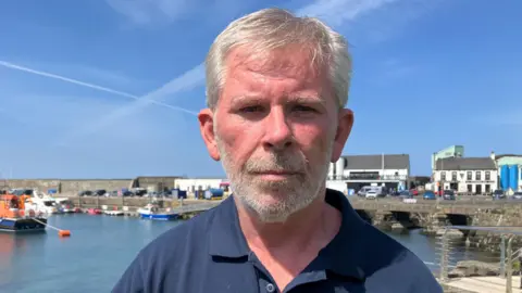 A man is standing in front of a small dock. He has grey hair and is wearing a navy polo shirt. In the background are a number of white buildings and one orange boat in the water. 