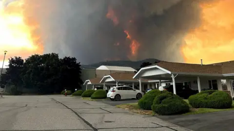 An image showing grey and orange smoke rising from behind a row of homes on a residential street in Summerland, British Columbia. 